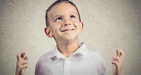 Junge schaut nach oben und hält beide Hände mit gekreuzten Fingern mit gebeugten Ellenbogen nach oben in Schulterhöhe.