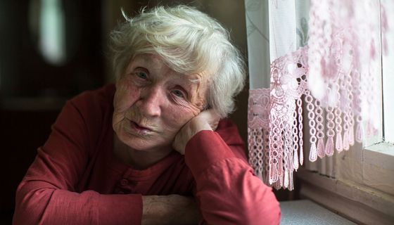 Alte Frau, stützt den Kopf auf ihre Hand und schaut in die Kamera.
