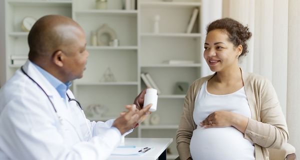 Eine schwangere Frau sitzt auf einem Stuhl mit einem Arzt im weißen Kittel. Er hält eine weise Dose in der Hand. Mit der anderen Hand deutet er auf die Dose.