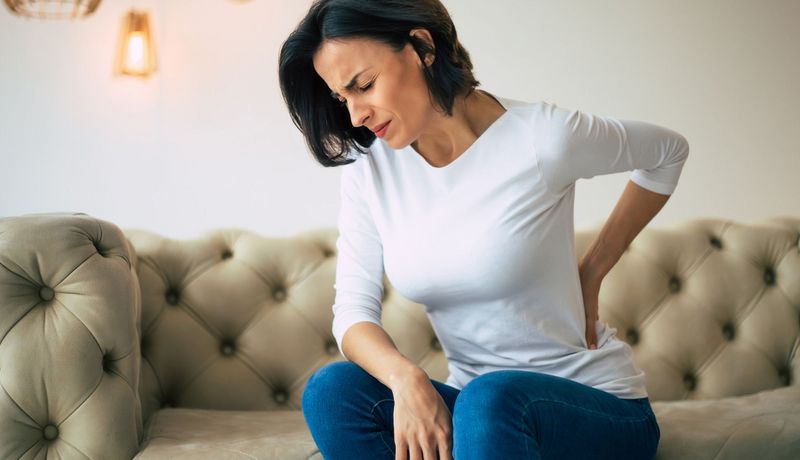 Frau sitzt auf dem Sofa und hält sich den Rücken, etwa auf Taillenhöhe.