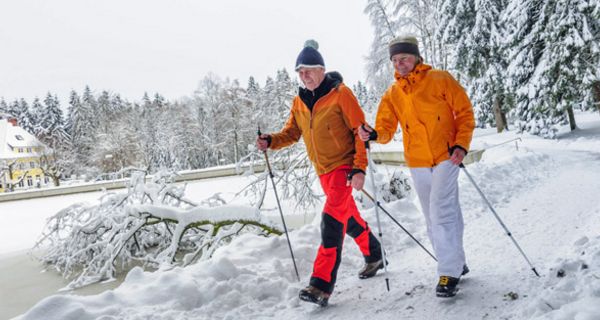 Senioren-Paar in Winterfunktionskleidung läuft mit Gehstöcken an einem zugefrorenen See in winterlicher Natur entlang