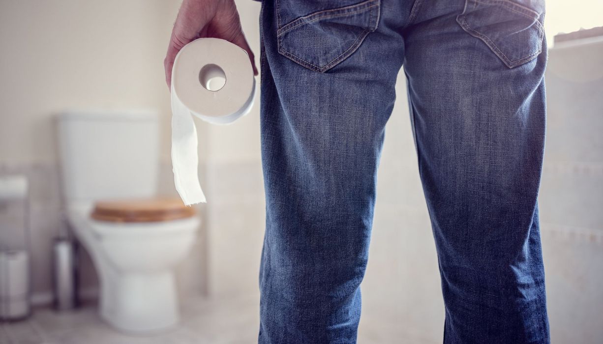Ein Mann hält eine Rolle Klopapier in der Hand und geht Richtung Toilette (unscharf im Hintergrund erkennbar).