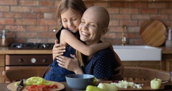 Krebspatientin umarmt ein Mädchen, vor ihnen liegt geschnittenes Gemüse.