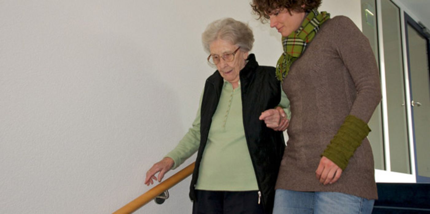 Seniorin wird von Frau um die 50 Treppe hinuntergeführt, hält sich mit einer Hand am Treppengeländer fest