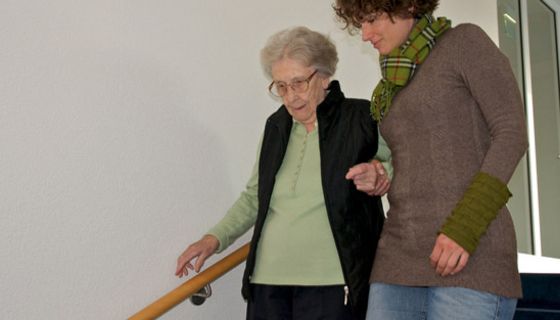 Pflegerin begleitet alte Frau die Treppe hinunter.