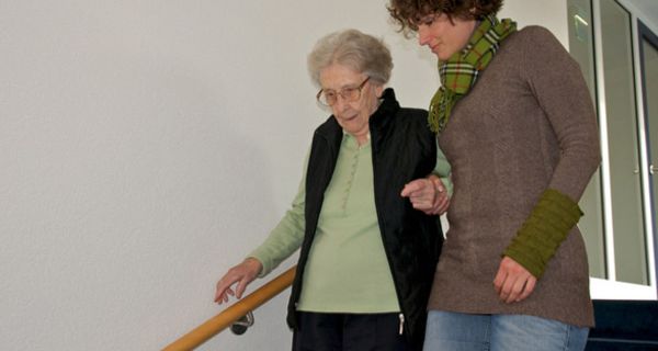 Seniorin wird von Frau um die 50 Treppe hinuntergeführt, hält sich mit einer Hand am Treppengeländer fest