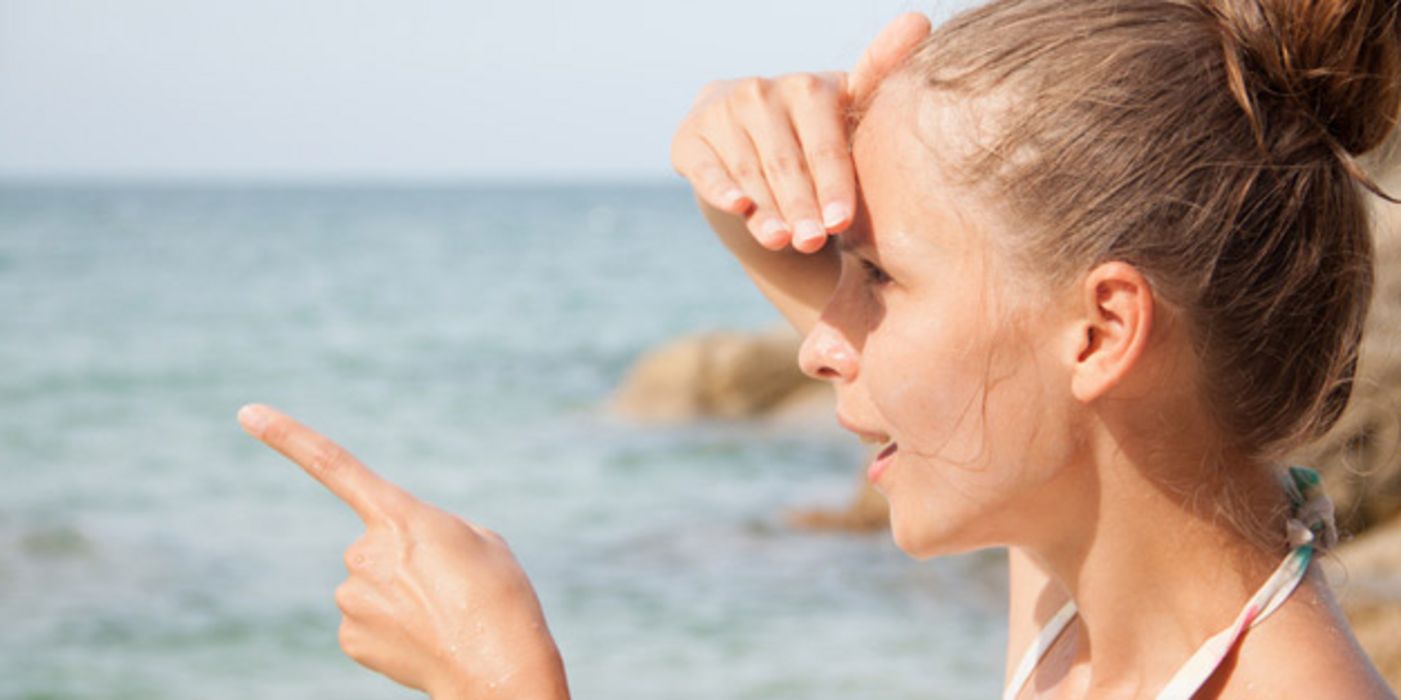 Junge Frau im Profil am Meer, hochgebundene Haare, schaut mit der einen Hand Augen vor der Sonne schützend, aufs Meer und deutet mit der anderen Hand in die Ferne