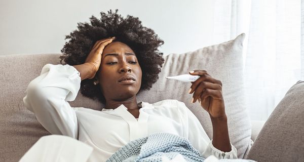 Eine junge Frau liegt in einer Decke auf dem Sofa, greift sich an den Kopf und blickt auf ein Fieberthermometer.
