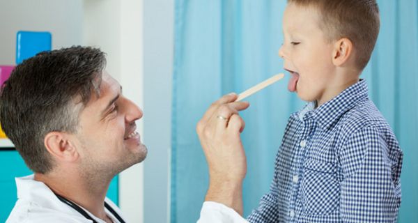 Freundlich lächelnder Arzt schaut Jungen (ca. 8 Jahre) in den Mund, Junge zeigt Zunge, die mit Spatel vom Arzt heruntergehalten wird