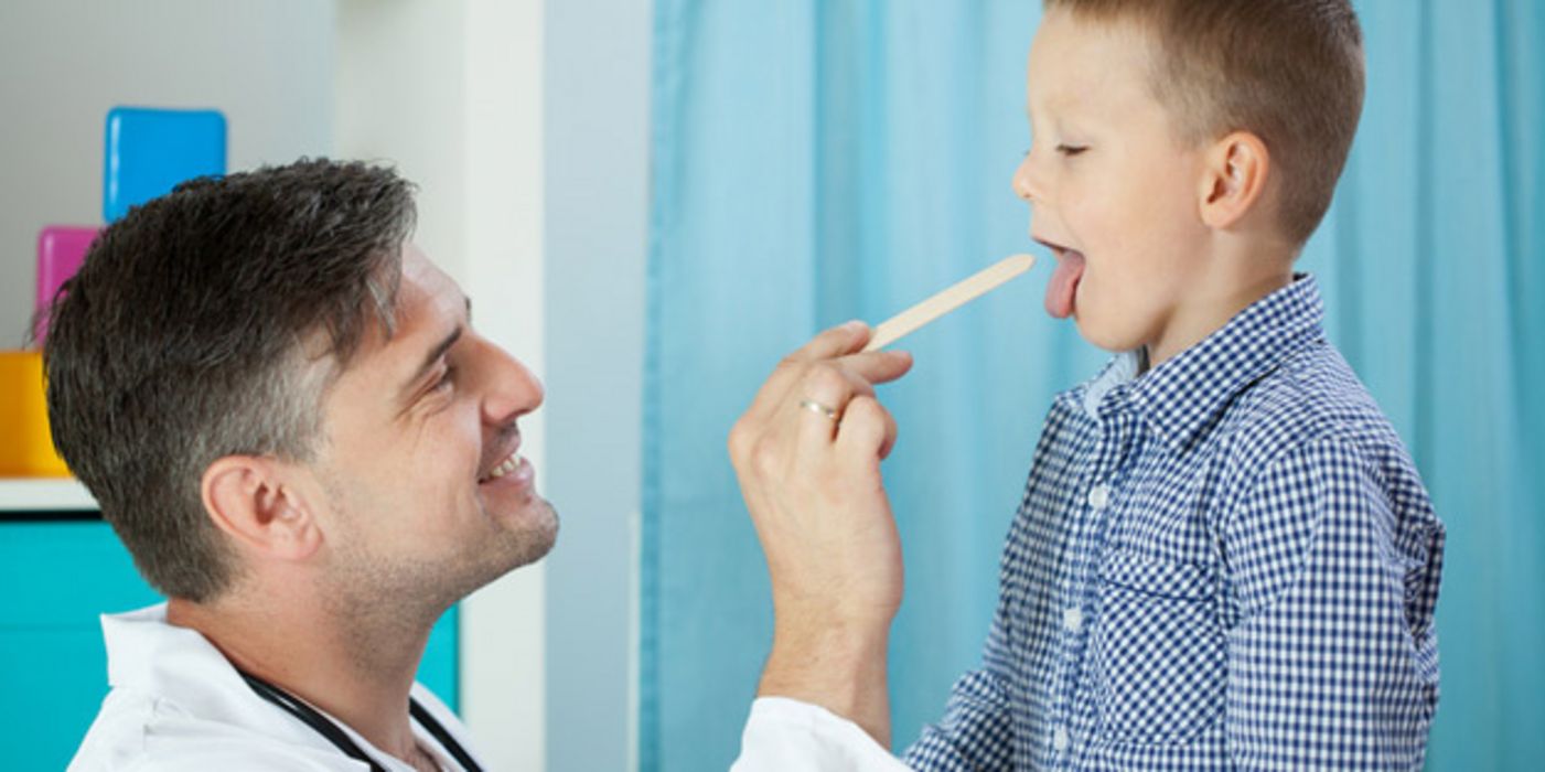 Freundlich lächelnder Arzt schaut Jungen (ca. 8 Jahre) in den Mund, Junge zeigt Zunge, die mit Spatel vom Arzt heruntergehalten wird