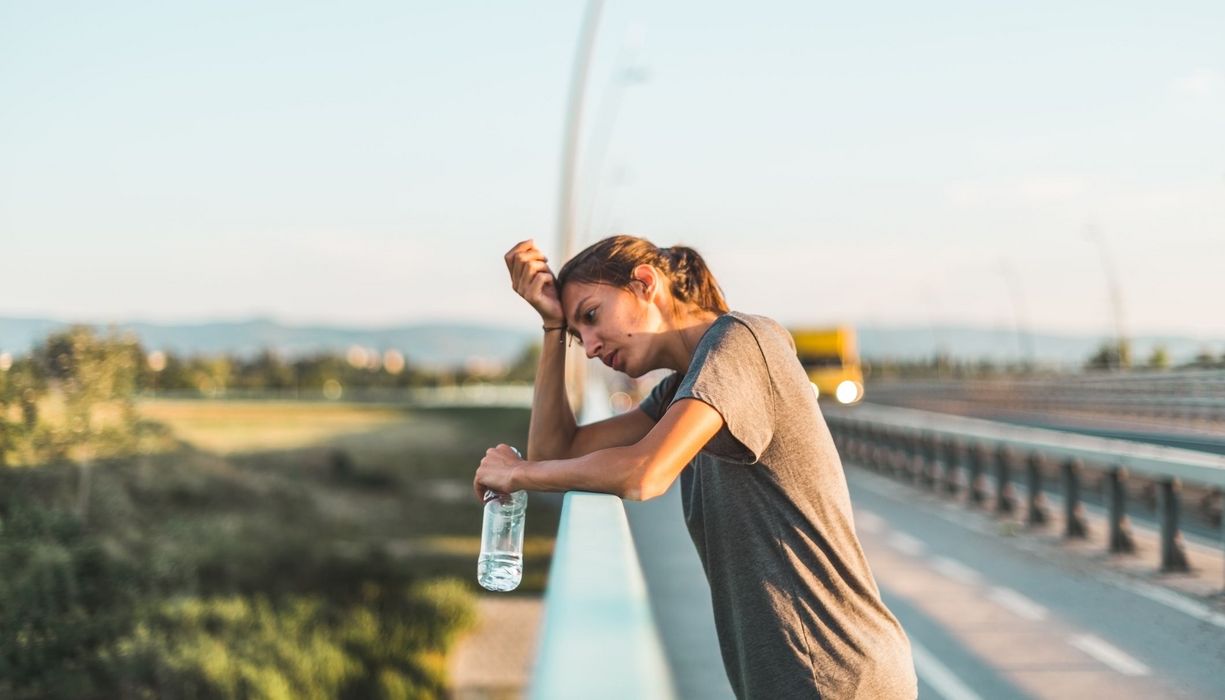 Frau in der Sommerhitze, lehnt an ein Geländer und trinkt Wasser.