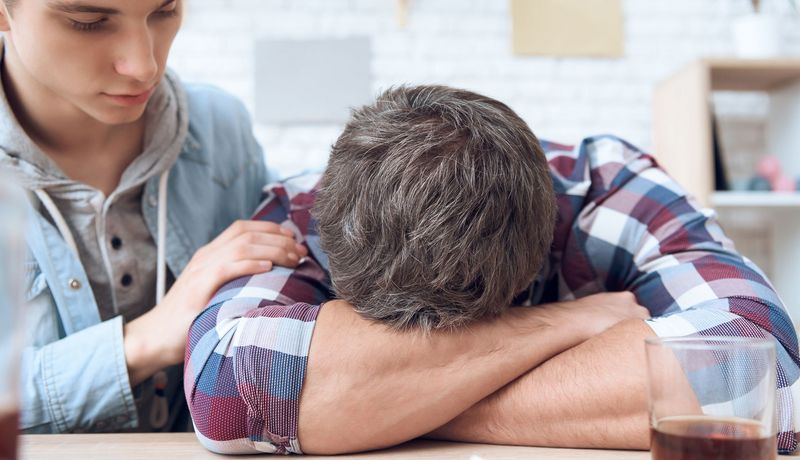 Mann liegt mit dem Kopf auf den Unterarmen auf einem Tisch, vor ihm steht ein Glas mit einer dunkelbraunen Flüssigkeit. Ein Teenager versucht seine Aufmerksamkeit zu bekommen.