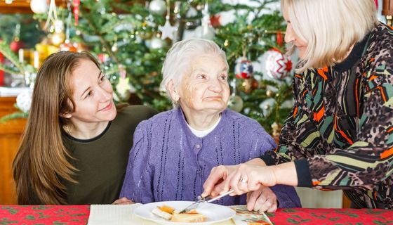 Eine ältere Dame sitzt an einem Tisch. Neben ihr ist eine lächelnde Junge Frau und eine zweite ältere Frau, die Essen auf dem Teller schneidet.