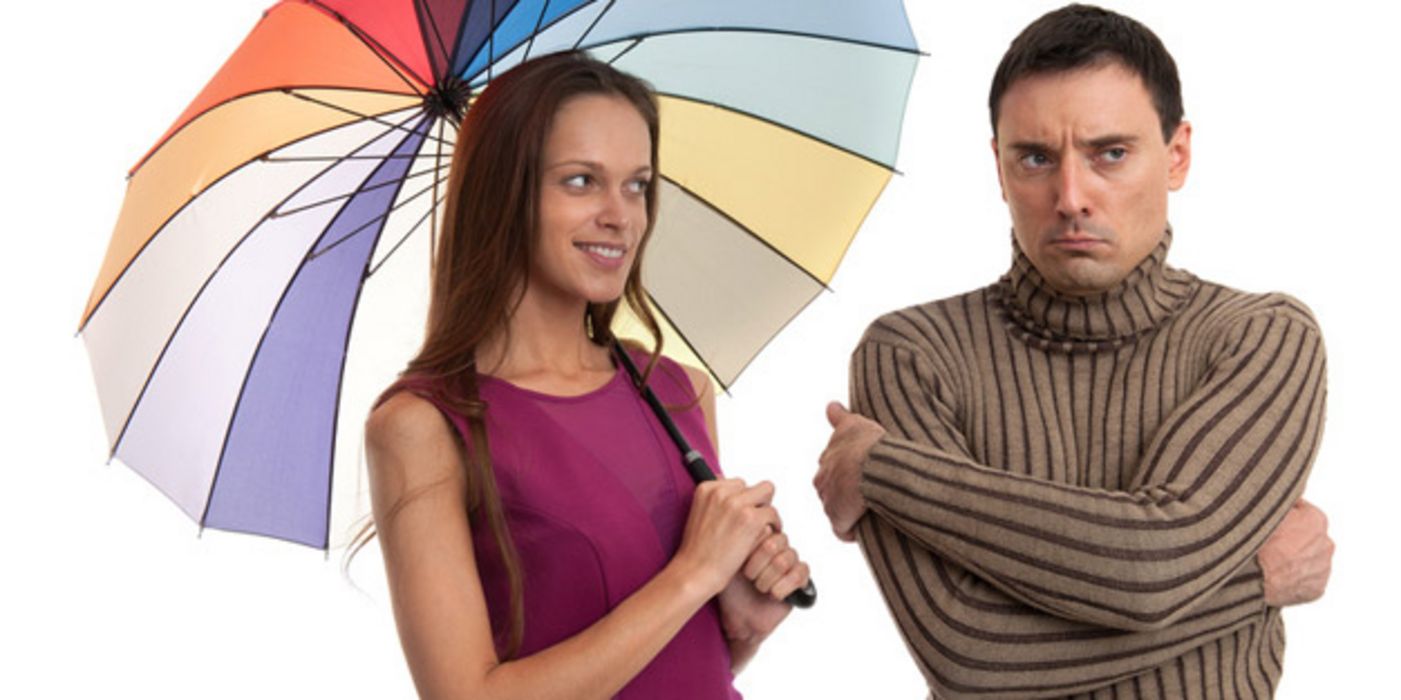 Fröhliche Frau mit buntem Regenschirm schaut auf einen Mann, der beleidigt guckt, eine Flunsch zieht und die Arme vor der Brust verschränkt hat