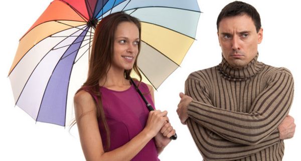 Fröhliche Frau mit buntem Regenschirm schaut auf einen Mann, der beleidigt guckt, eine Flunsch zieht und die Arme vor der Brust verschränkt hat
