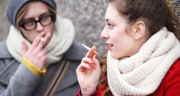 Junge rauchende Frau im Vordergrund, im Hintergrund junger, rauchender Mann, beide in Winterkleidung mit Mützen und Schals, Frau im Halbprofil, Mann frontal