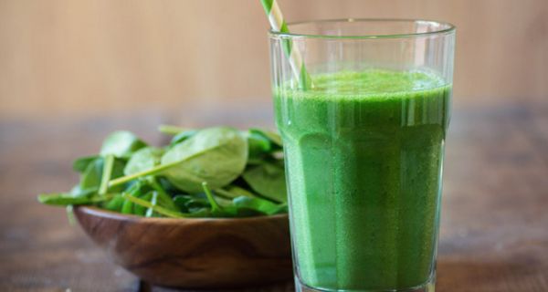 Spinatdrink in hohem Glas mit weiß-grünem Strohhalm auf dunkelm Holztisch, links etwas dahinter dunkles Holzschälchen mit jungen Spinatblättern