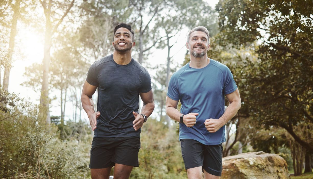 Ein älterer, beginnend grauhaariger Mann im blauen Shirt joggt mit einem dunkelhäutigen Mann in schwarzem Shirt durch einen Park.