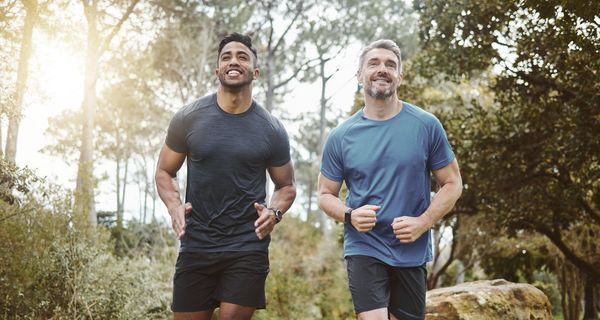Ein älterer, beginnend grauhaariger Mann im blauen Shirt joggt mit einem dunkelhäutigen Mann in schwarzem Shirt durch einen Park.