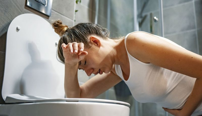 Frau beugt sich über eine Toilettenschüssel und hält sich den Bauch. Sie sieht nicht glücklich aus.