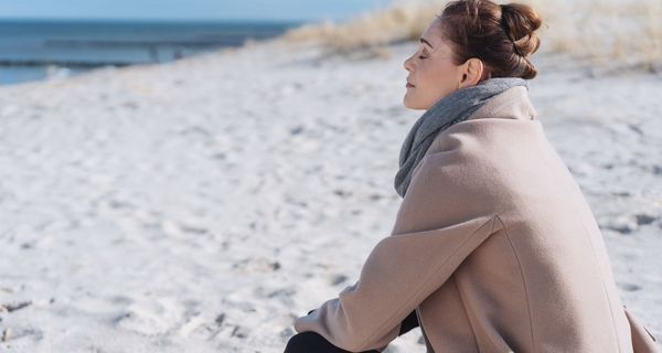 Junge Frau, sitzt an einem kühleren Tag warm angezogen am Strand und sonnt sich.