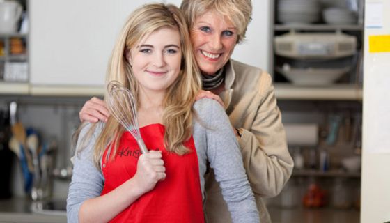 Küchenszene: Teenagertochter im Vordergrund, blonde Haare, rote Schürze, Schneebesen in der rechten Hand, dahinter sie an den Schultern haltend Mutter, beide in die Kamera lächelnd