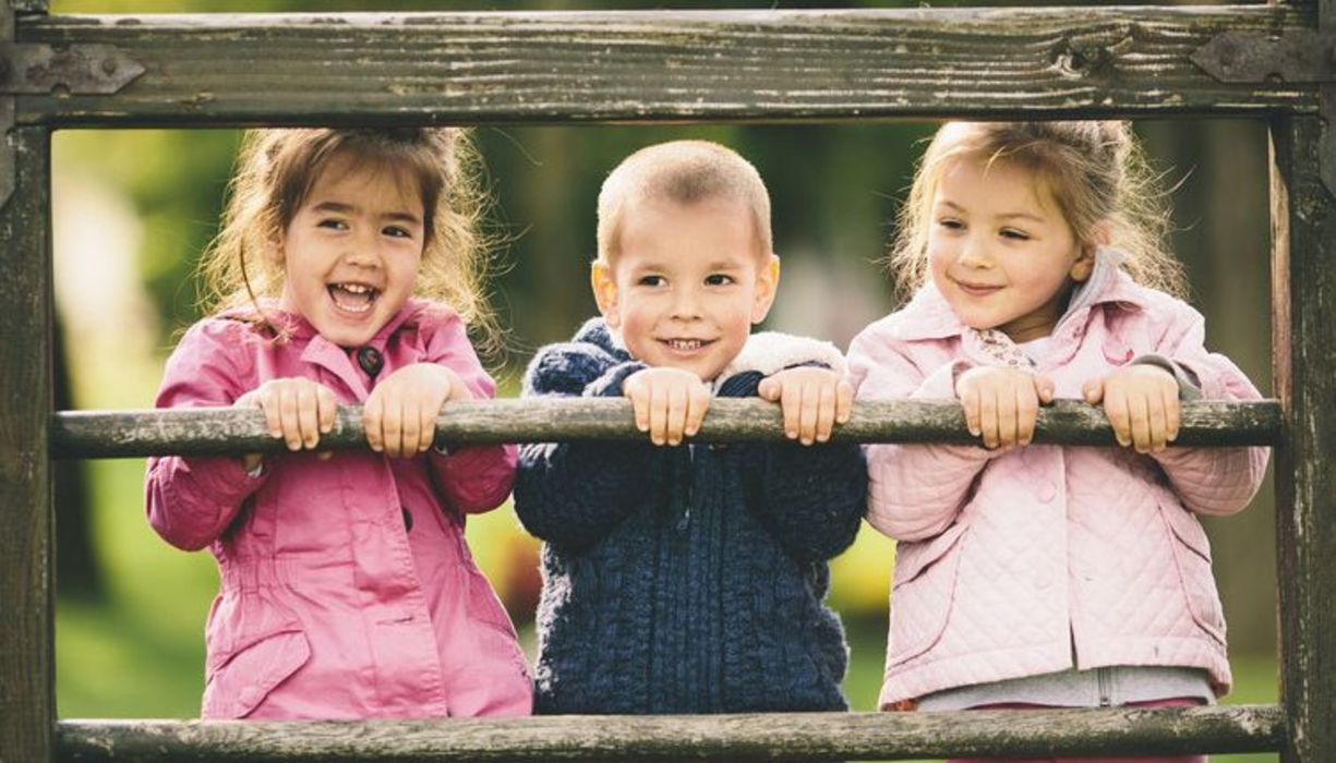 Drei vergnügte Kinder, ca. fünfjährig, rechts und links Mädchen, Junge in der Mitte, auf einem Holzzaun stehend und sich mit den Händen festhaltend