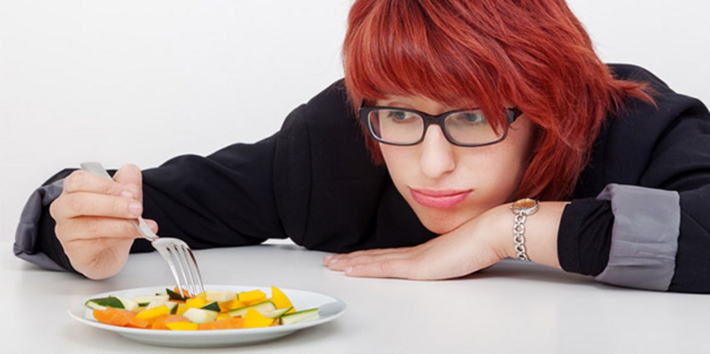 Junge rothaarige Frau, schwarzrandige Brille, guckt missmutig über Tischkante auf Teller mit geschnittenem Obst
