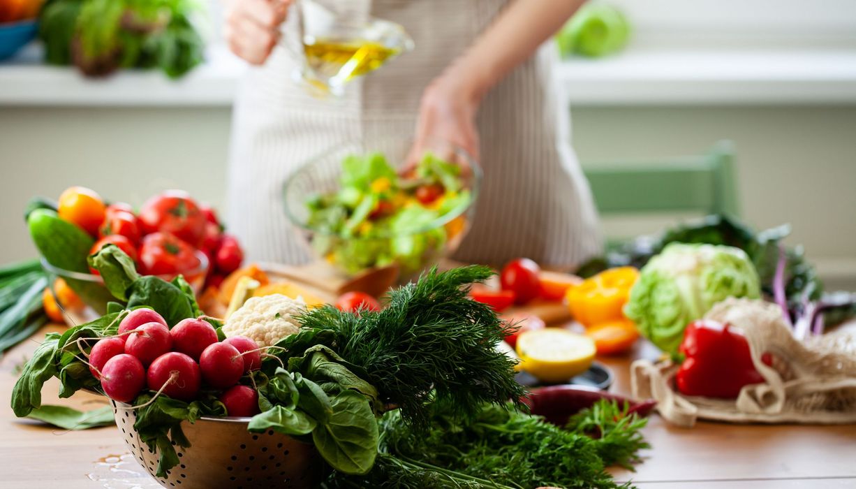 Im Vordergrund: Verschiedene Gemüsesorten wie Radiesschen, Karotten, Salat, Paprika und Zwiebel. Im Hintergrund: Eine Person bereitet einen Salat zu und fügt etwas Öl hinzu. 