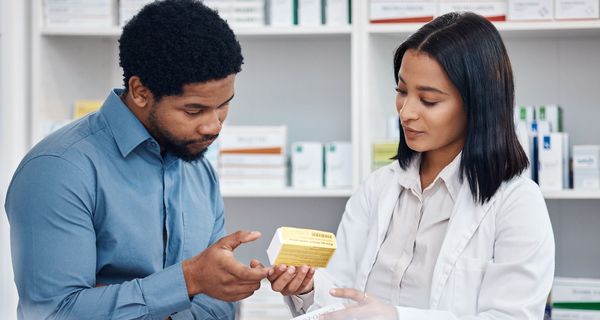 Ein dunkelhäutiger Mann steht neben einer Apothekerin im weißen Kittel und lässt sich beraten.
