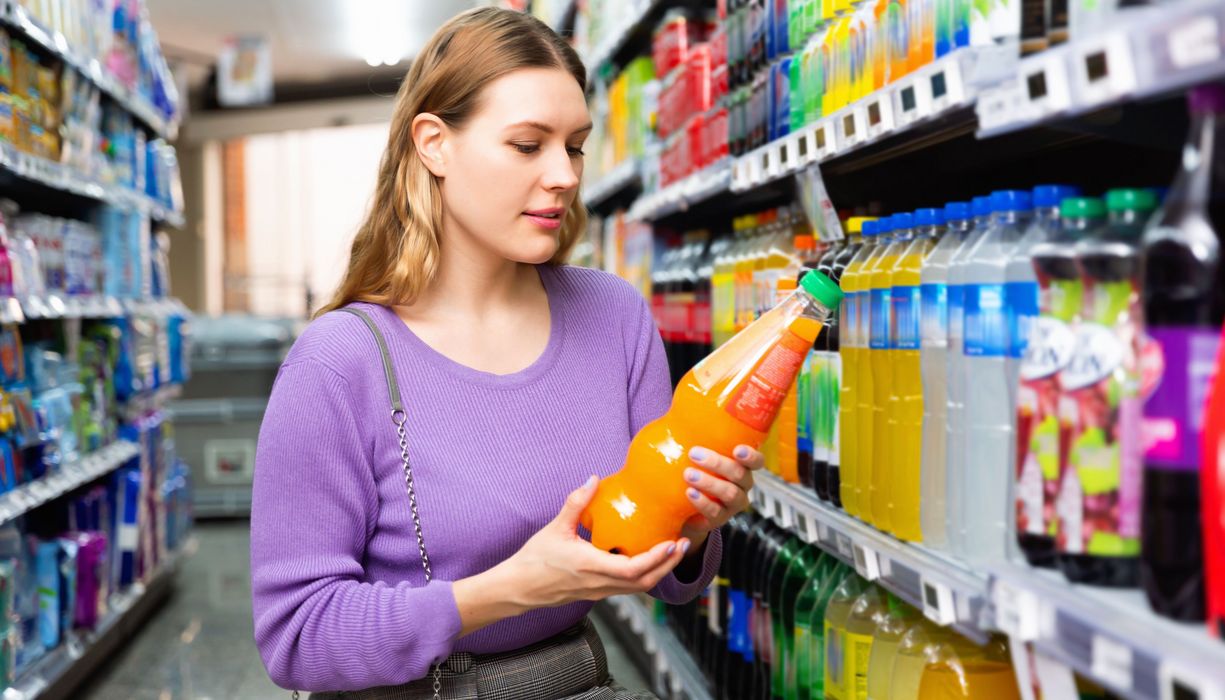 Eine Frau steht vor dem Getränkeregal und schaut auf die Zutatenliste eines Softdrinks.