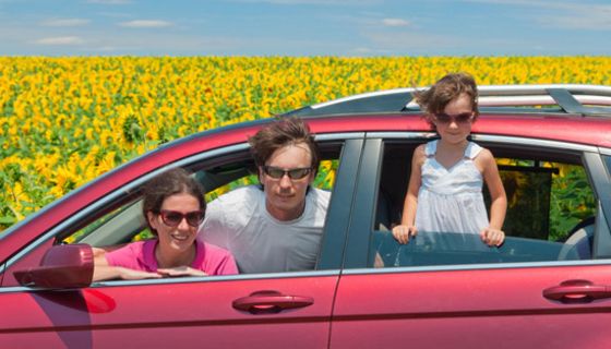 Mutter, Vater, Tochter schauen lachend aus Fenstern eines roten Autos, im Hintergrund Sonnenblumen und blauer Himmel