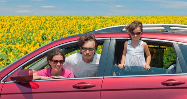 Mutter, Vater, Tochter schauen lachend aus Fenstern eines roten Autos, im Hintergrund Sonnenblumen und blauer Himmel