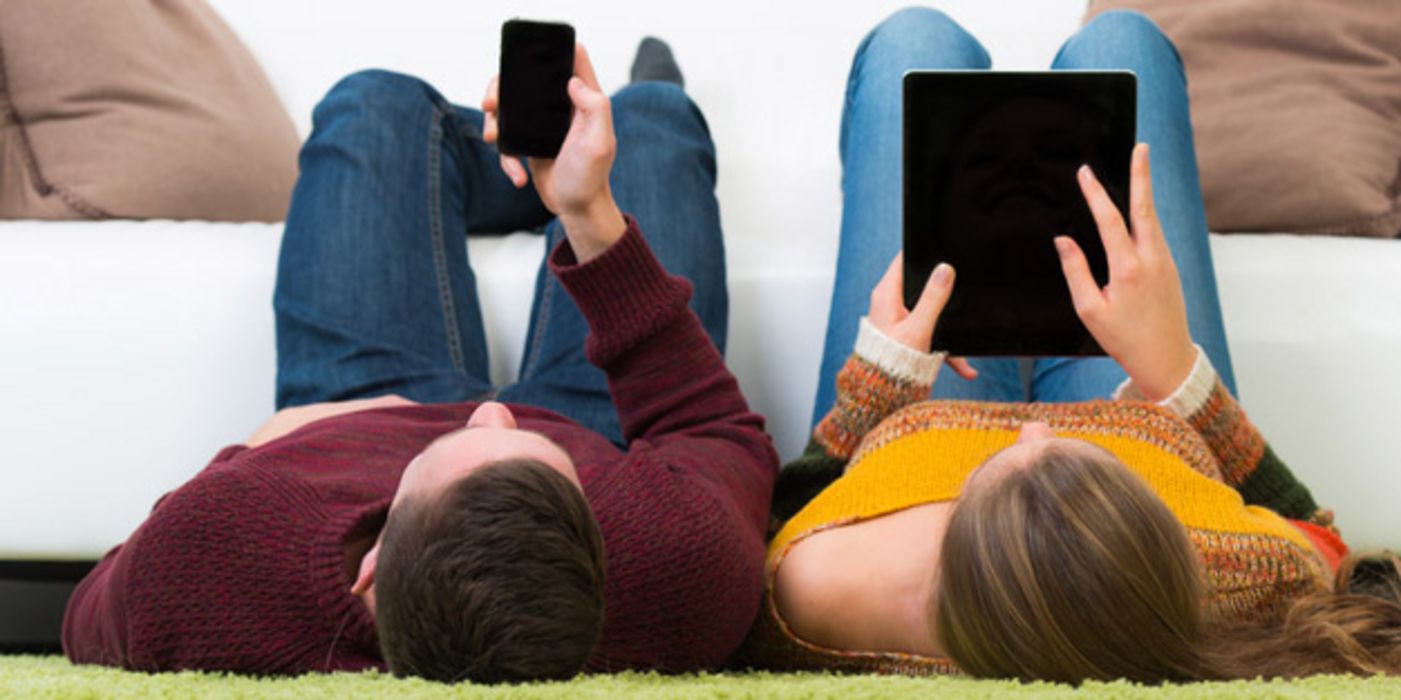 Zwei Jugendliche liegen auf dem Rücken und lesen auf Tablet und Smartphone.