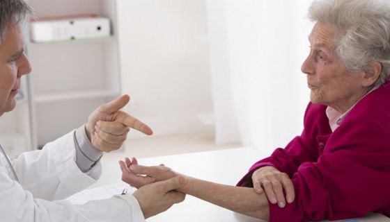 Arzt (linker Bildrand) erklärt Seniorin (rechts) etwas, dabei betrachtet und hält ihre Hand