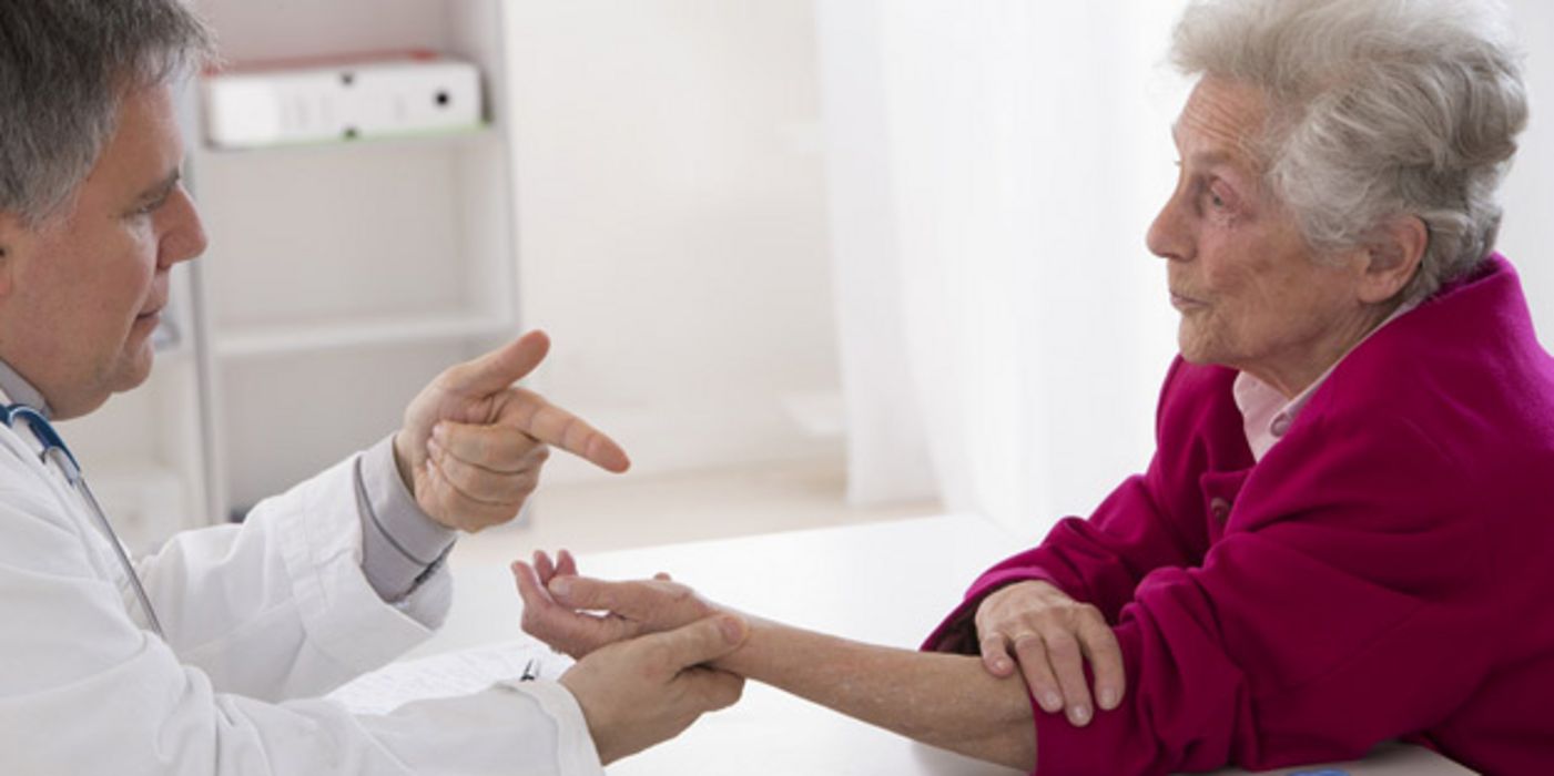 Arzt (linker Bildrand) erklärt Seniorin (rechts) etwas, dabei betrachtet und hält ihre Hand