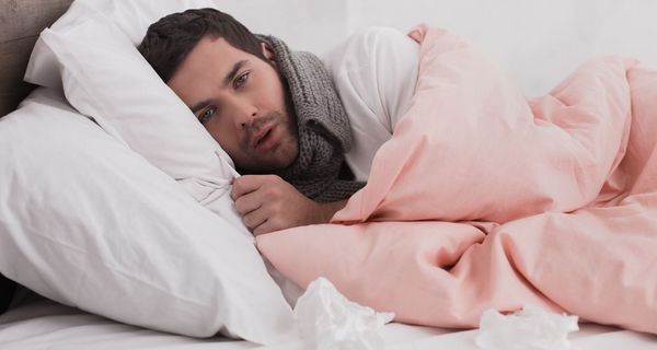 Junger Mann liegt im Bett mit Schal um den Hals, ein Fieberthermometer in der Hand. Vor ihm: Medikamente und Taschentücher.