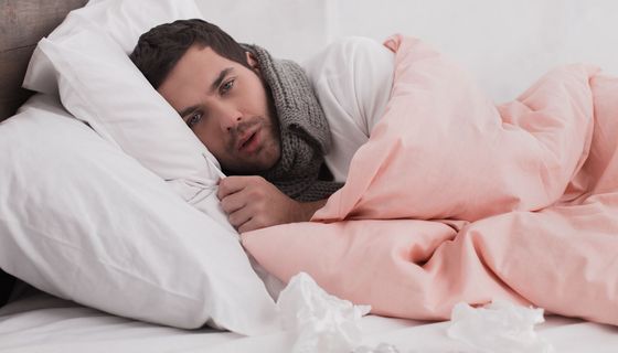 Junger Mann liegt im Bett mit Schal um den Hals, ein Fieberthermometer in der Hand. Vor ihm: Medikamente und Taschentücher.