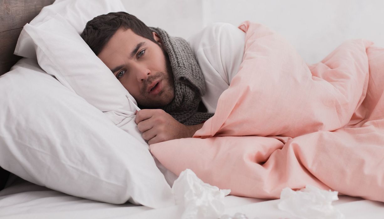Junger Mann liegt im Bett mit Schal um den Hals, ein Fieberthermometer in der Hand. Vor ihm: Medikamente und Taschentücher.