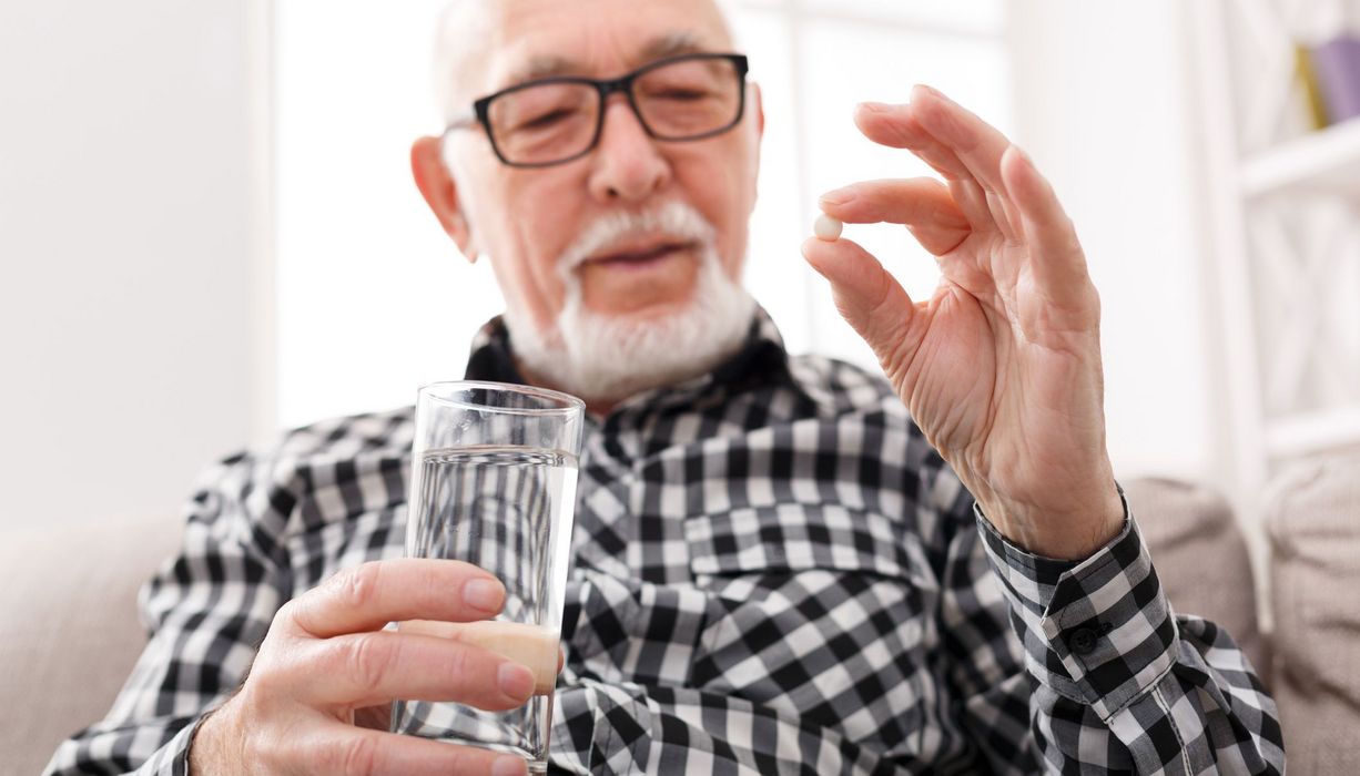Älterer Mann hält ein Glas Wasser in der Hand und nimmt eine Tablette ein 