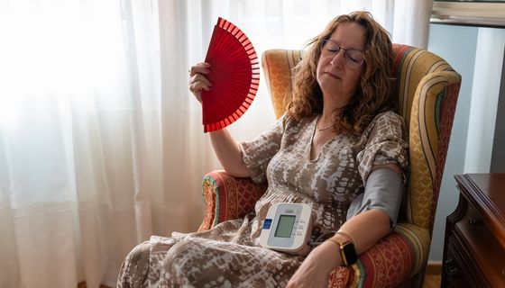 Dame sitzt im Sessel, in der Hand einen Fächer mit Blutdruckmessgerät auf dem Schoß und Manschette am Arm.