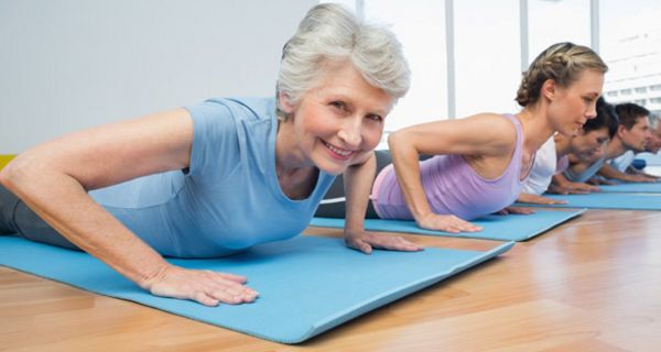 Gruppe beim Yoga auf Matten auf dem Bauch (Kobra), Seniorin im Vordergrund lächelt in die Kamera.