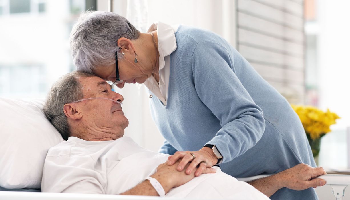 Mann im Krankenhausbett, seine Frau beugt sich über ihn.