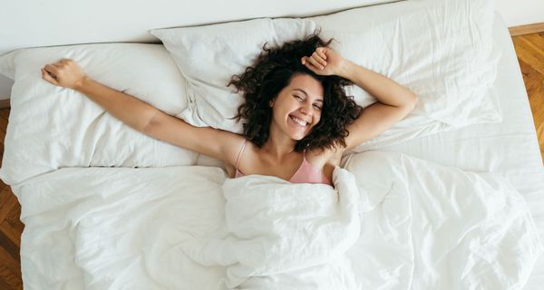 Eine junge Frau liegt im Bett. Sie ist wach, lächelt und streckt ihre Arme aus.