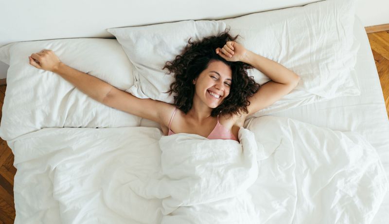 Eine junge Frau liegt im Bett. Sie ist wach, lächelt und streckt ihre Arme aus.