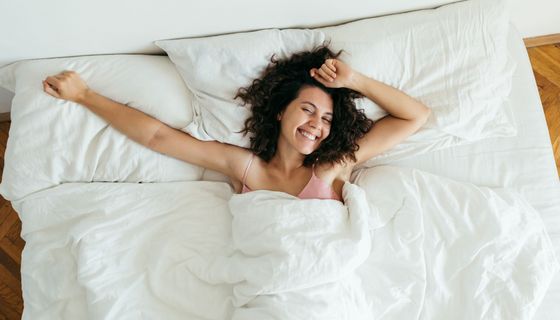 Eine junge Frau liegt im Bett. Sie ist wach, lächelt und streckt ihre Arme aus.