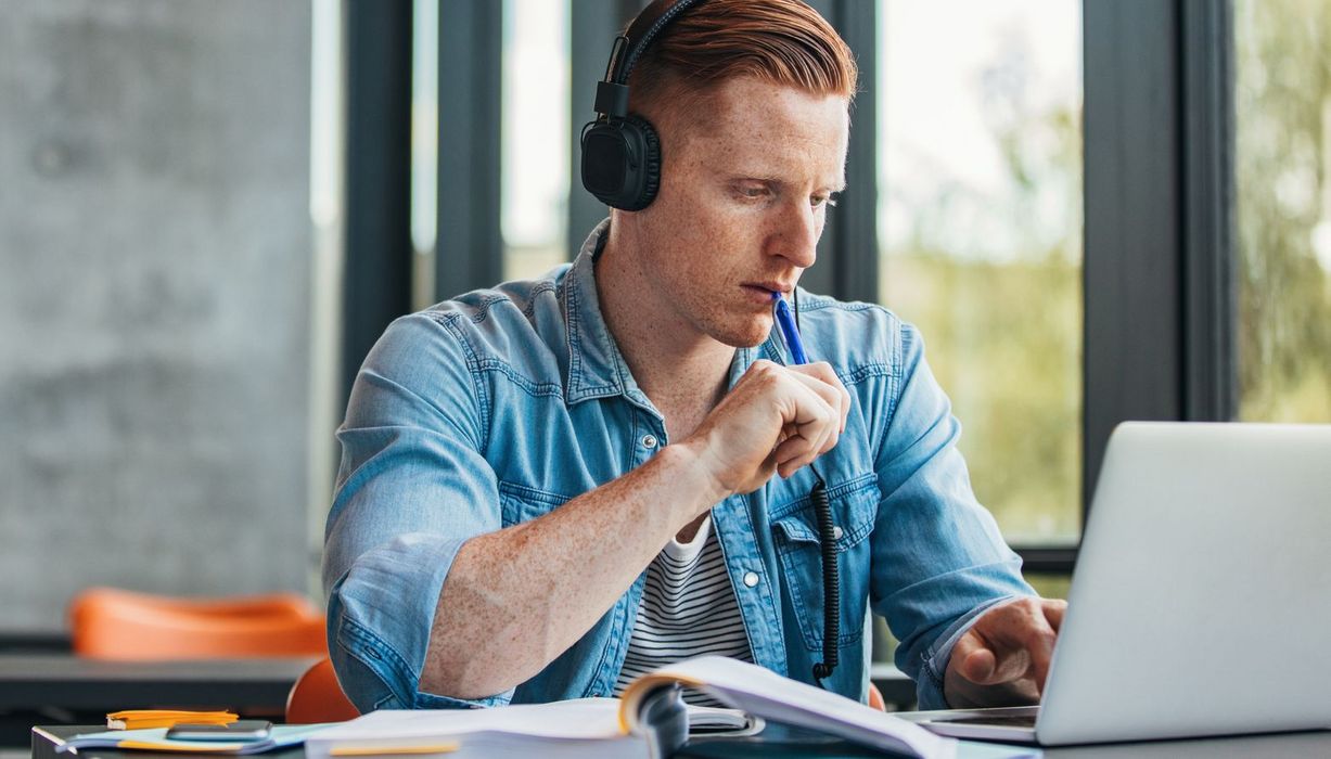 Junger Mann mit Kopfhörern sitzt vor einem aufgeschlagenen Buch und einem Laptop. Er tippt mit dem Stift an seine Unterlippe.