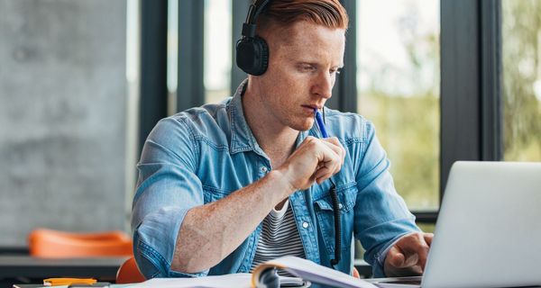 Junger Mann mit Kopfhörern sitzt vor einem aufgeschlagenen Buch und einem Laptop. Er tippt mit dem Stift an seine Unterlippe.
