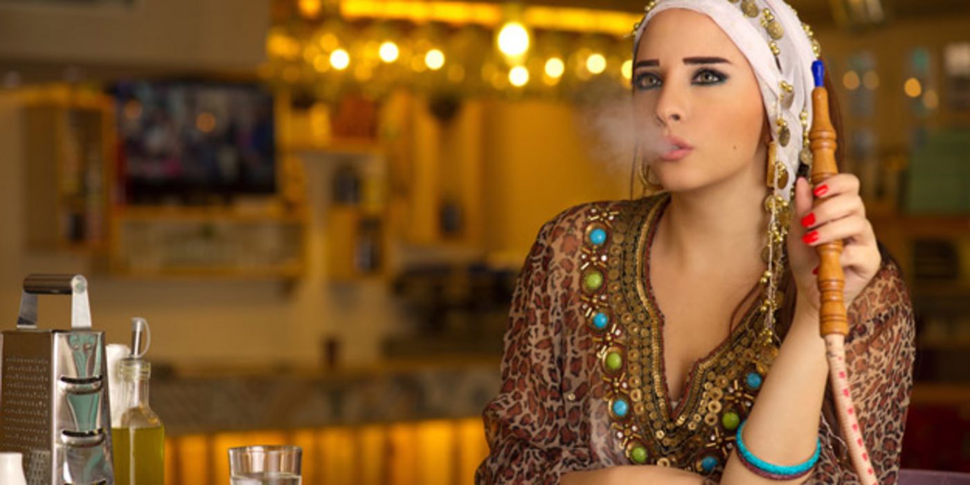 Junge Frau, modern folkloristisch gekleidet an einem Tisch sitzend raucht eine Shisha