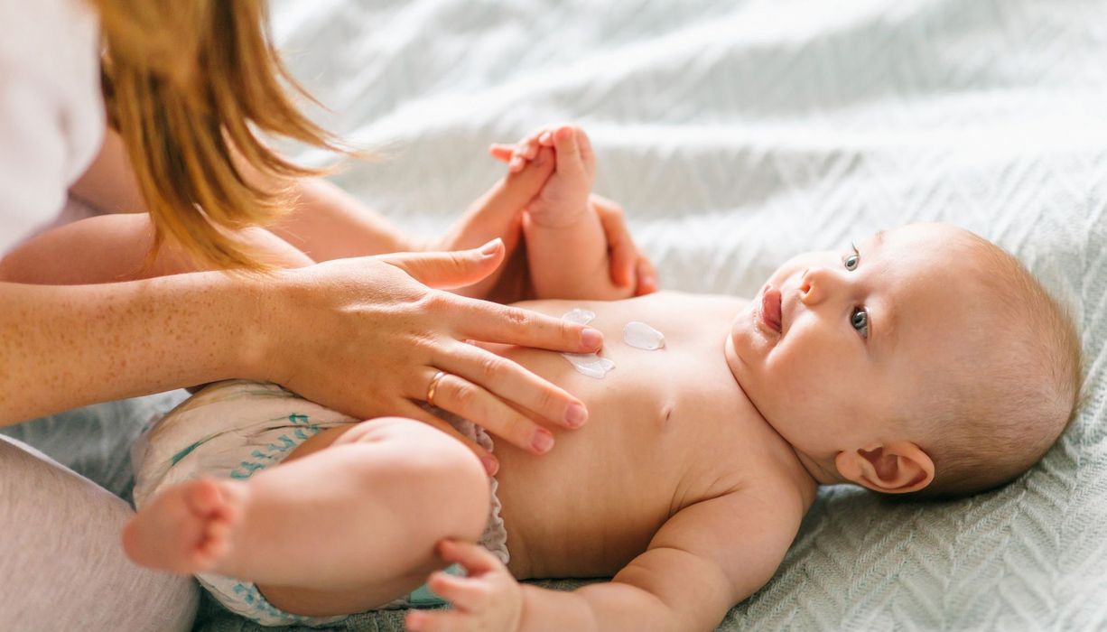 Babys wird eingecremt. Hände und Haare einer Frau sind zu sehen.